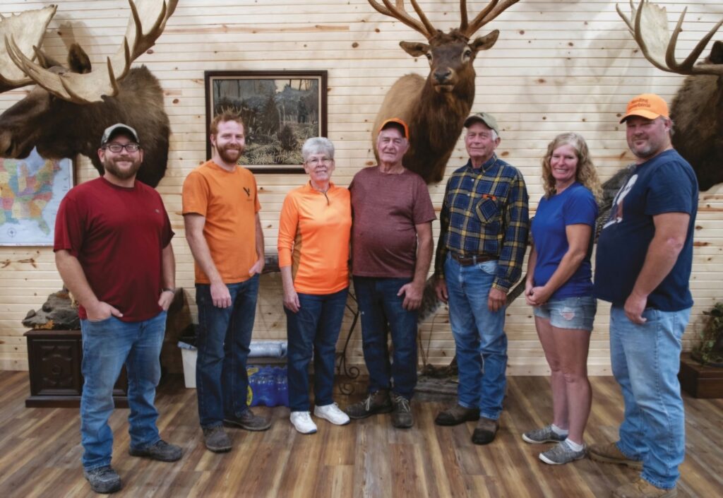 Seven people standing indoors in front of a wall with mounted elk heads. They are casually dressed, some in hats, in a wood-paneled room.